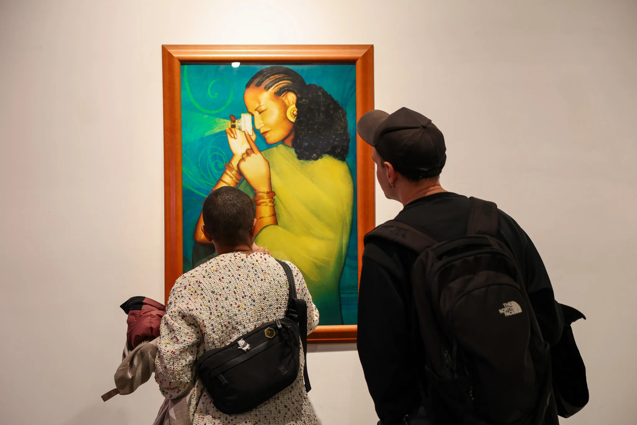 Two visitors view a framed painting of a woman applying gold light to her forehead at Future Legends