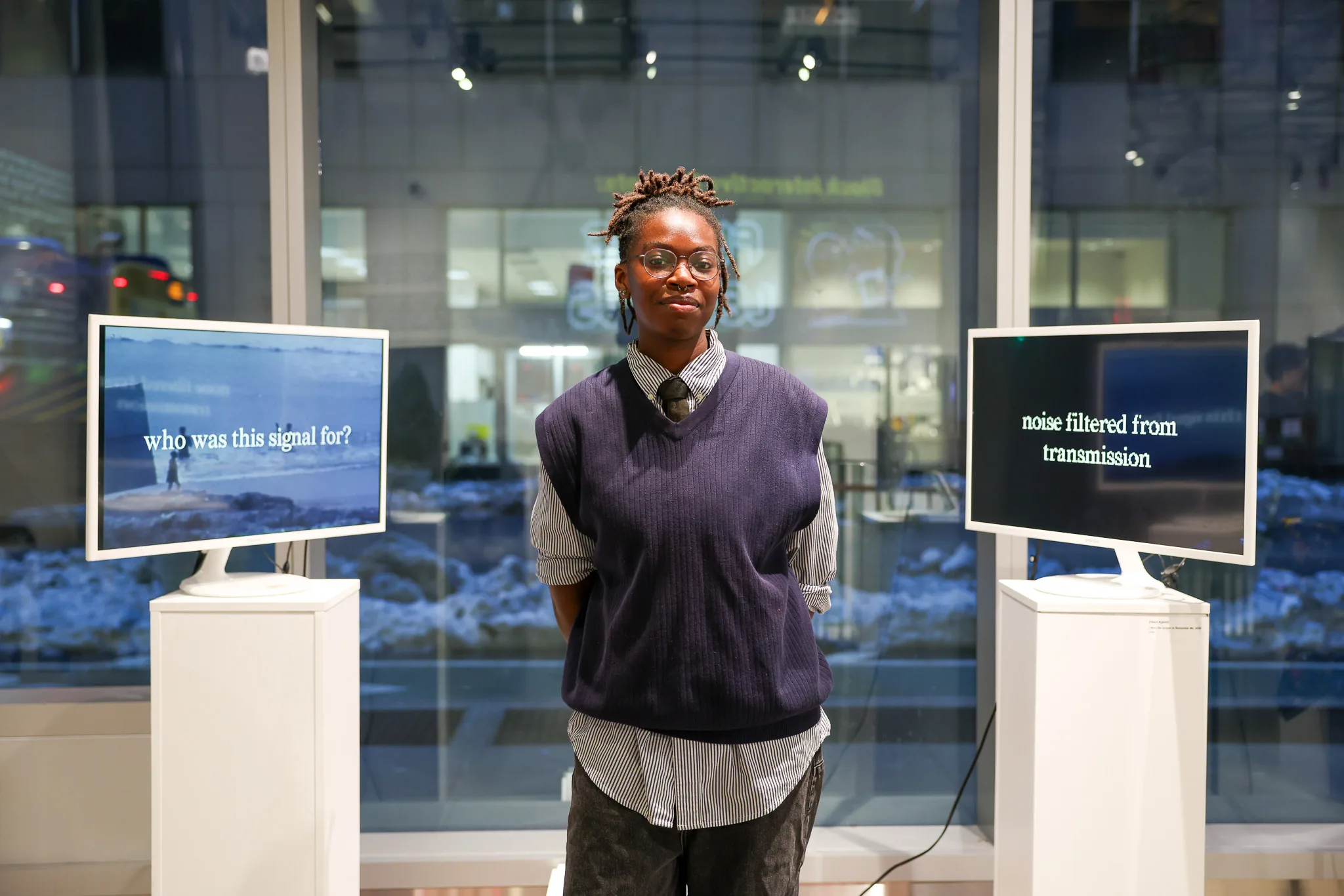An artist stands between two video monitors reading 'who was this signal for?' and 'noise filtered from transmission'
